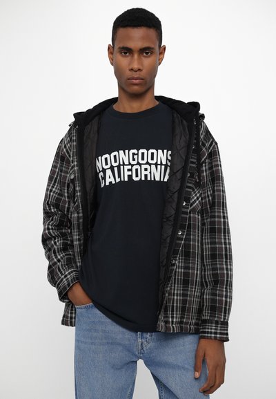 Veste en tartan noir et gris avec doublure matelassée noire, superposée à un t-shirt sombre portant "NOONGOONS CALIFORNIA" en texte blanc, jeans en denim clair.