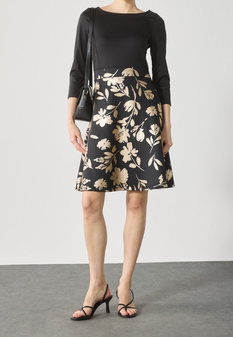 Femme portant un haut noir à manches trois-quarts et une jupe noire longueur genou avec un motif floral beige, tenant un sac à main noir et des talons noirs.