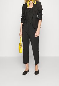 Blazer noir sur un haut noir, associé à un pantalon noir. Comprend un sac à main jaune et une écharpe à motifs jaunes. Chaussures plates noires.