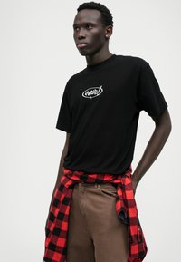 Schwarzes Baumwoll-T-Shirt mit einem weißen Logo; kombiniert mit braunen Hosen und einem rot-schwarzen karierten Hemd, das um die Taille gebunden ist.