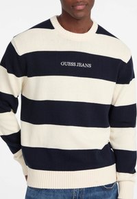 Gestreifter Strickpullover in Marineblau und Creme mit gerippten Bündchen und Saum, der in der Mitte mit "GUESS JEANS" bestickt ist. Standard Crew-Neck-Design.