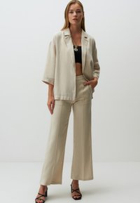 Blazer en lin beige avec une coupe décontractée, associé à un pantalon large assorti. Top court noir en dessous ; le mannequin porte des talons aiguilles noirs.