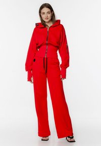 Czerwona bluza z kapturem i zamkiem, z dużymi rękawami i kieszenią z przodu, w zestawie z dopasowanymi spodniami o szerokich nogawkach, wykonanymi z miękkiego materiału.