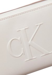 Hellbeige Lederbeutel mit geprägt "CK"-Logo. Glatte Textur mit einem Metallreißverschluss. Minimalistisches Design, rechteckige Form.