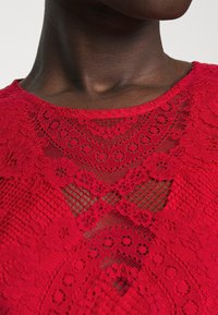 Gros plan d'une personne portant un haut en dentelle rouge avec des motifs floraux et des détails en maille géométrique autour du col.