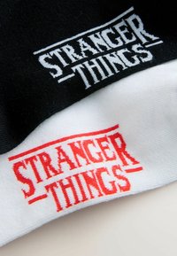 To huehuer er stablet, en sort med hvid "STRANGER THINGS"-logo og en hvid med rød "STRANGER THINGS"-logo, begge i strikket stof.