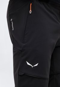 Τμήμα μαύρου αθλητικού παντελονιού με πορτοκαλί τράβηγμα φερμουάρ, πλαϊνή τσέπη, κείμενο "Durastretch" και λευκό γωνιακό λογότυπο κοντά στη ραφή του μηρού.