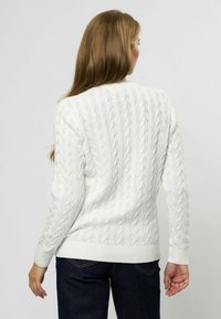 Fehér, kötött cable-knit pulóver bordás mandzsettával és szegéllyel. Függőleges irányú texturált minta. Sötét farmernadrággal viselve.