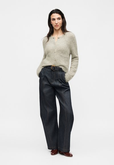 Femme aux cheveux foncés portant un cardigan beige duveteux boutonné, un jean large taille haute foncé avec ceinture, et des chaussures plates marron, debout les mains dans les poches.