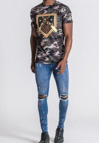 Kamouflagemönstrad t-shirt med guldtrycket "GK Army", matchad med slitna blå jeans som har färgprickar och knäskärningar, bärs med svarta sneakers.