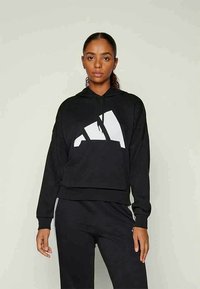 Mulher com cabelo escuro preso, vestindo um hoodie preto com um logotipo geométrico branco e calças pretas com riscas brancas, de pé contra um fundo simples.