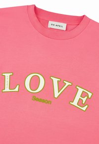 Růžové bavlněné tričko s výrazným zeleným nápisem „LOVE“ a menším bílým nápisem „Season“. Design s kulatým výstřihem, hladká textura. Štítek: „OH APRIL.“