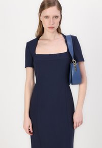 Morska obleka s kvadratnim izrezom in kratkimi rokavi, prilegajoča se in gladke teksture. Modra torbica z naramnico visi z rame in ima minimalistične detajle.