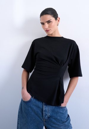 Joven con el cabello oscuro recogido hacia atrás, lleva una camiseta negra de manga corta con detalles asimétricos y vaqueros azules holgados, manos en los bolsillos.