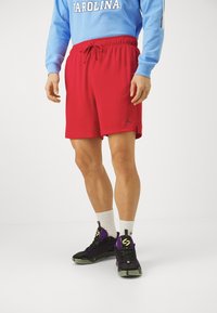 Punased spordilühikesed püksid musta logoga, elastse vöökoha ja stringiga. Kombineeritud mitmevärviliste tossudega, valgete sokkidega ja sinise kapuutsiga dressipluusiga.