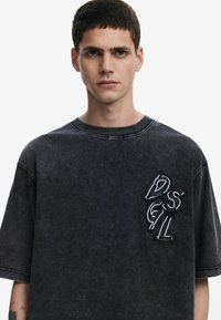 Camiseta de manga corta de mezclilla negra con un parche de logo texturizado en el lado izquierdo del pecho y costuras en contraste a lo largo del escote.
