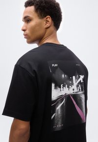 Νέος άνδρας με μαύρο t-shirt που έχει ασπρόμαυρη εκτύπωση δρόμου πόλης και τη λέξη "PLAY" στην πλάτη, στραμμένος στο πλάι μπροστά σε απλό φόντο.