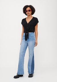 Top nera a maniche corte con bottoni e fiocco frontale, abbinata a jeans flare azzurri chiari con pannello laterale a contrasto, completata da stivali neri.