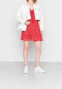 Abito rosso con fiori e orlo arricciato, indossato sotto una giacca di jeans bianca corta. Abbinato a sneakers bianche e una piccola borsa bianca.