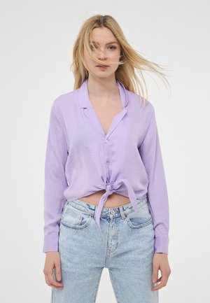 Camicia button-up lavanda con design a cravatta frontale, maniche lunghe e vestibilità comoda, abbinata a jeans in denim chiaro.