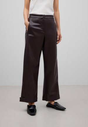 Femme portant un pantalon en cuir marron foncé à jambes larges avec des ourlets retournés, associé à des mocassins noirs à enfiler et un haut blanc sans manches.