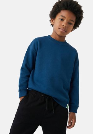 Enfant aux cheveux bouclés portant un sweat-shirt bleu et un pantalon noir, debout avec une main dans la poche sur un fond uni.