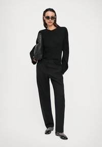 Zwarte gebreide trui met een gestructureerd patroon, losse broek en platte schoenen. Accessoires omvatten een zwarte tas en ronde zonnebril.