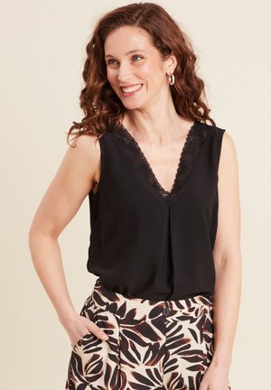 Smiling woman with curly brown hair wears sleeveless black top with lace V-neck and patterned beige and black pants, hand in pocket.