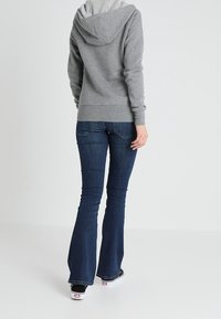 Persona con una felpa grigia, jeans a zampa di elefante blu scuro e sneaker nere, in piedi di spalle contro uno sfondo chiaro e semplice.