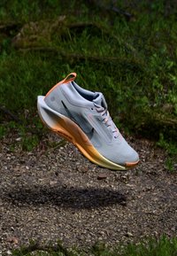 Pantofii sport de culoare gri deschis, cu accente portocalii și un superior texturat. Prezintă un design elegant, talpă căptușită și branding negru.