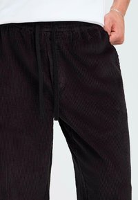 Schwarze Corduroy-Hosen mit einem strukturierten Rippenmuster, ausgestattet mit einem elastischen Bund und einem Kordelzug. Sichtbare Seitentasche auf der linken Seite.