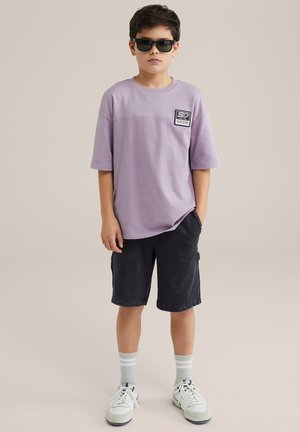 Camiseta oversized lila con un parche en el pecho, combinada con pantalones cortos azul marino. Zapatillas blancas con calcetines grises a rayas. Gafas de sol negras.