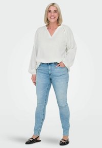 Bílá volná blůza s výstřihem do V a texturovaným detailem na rukávech; spárovaná se světle modrými skinny džíny a černými slip-on botami.