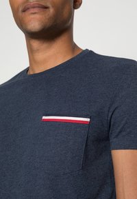 Mörkblå t-shirt med korta ärmar och en texturerad ficka med röd, vit och blå randig detalj. Klassisk rundhalsad design.