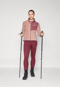Giacca in pile fucsia e bordeaux con zip completa, abbinata a leggings bordeaux aderenti. Scarpe da trekking nere e bastoni. Design minimalista.
