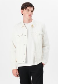 Hvit denimjakke med to brystlommer og knappelukking. Båret over en enkel hvit skjorte, kombinert med svarte bukser. Minimalistisk design.