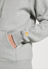 Harmaa huppari, jossa on pehmeä pinta, kangaroo-tasku ja pieni keltainen brodeerattu logo hihassa.