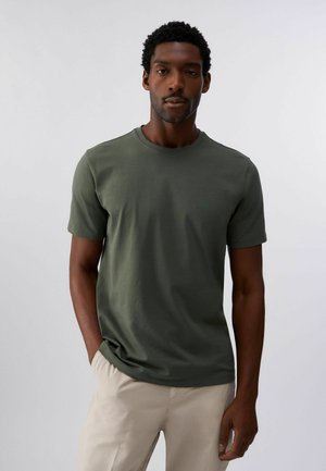 Mężczyzna z krótkimi kręconymi włosami, ubrany w prostą oliwkowozieloną koszulkę i beżowe spodnie, stoi z jedną ręką w kieszeni na białym tle.