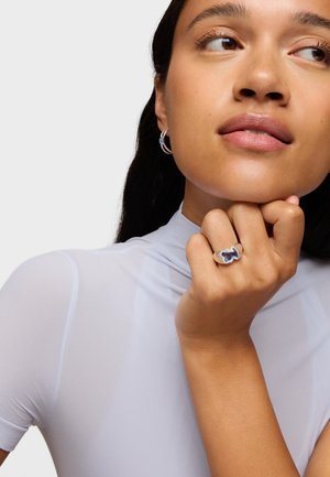 Anillo de plata con un diseño floral intrincado en azul oscuro, llevado en una mano de tono de piel claro. La modelo tiene el cabello largo y oscuro y lleva una blusa clara.