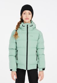Hellgrüne Pufferjacke mit Reißverschluss, verstellbare Kapuze und schwarzen Akzenten. Schwarze gerippte Strickmütze auf dem Kopf getragen.