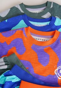 Trzy bluzy crewneck ułożone w stosie: zielony kamuflaż, niebieska z turkusowymi akcentami oraz pomarańczowo-fioletowy wzór abstrakcyjny z białymi detalami graficznymi.