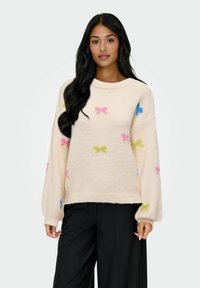 Vrouw met lang zwart haar draagt een roomkleurige trui met kleurrijke vlinderpatronen en zwarte wijde broek, staande tegen een platte achtergrond.