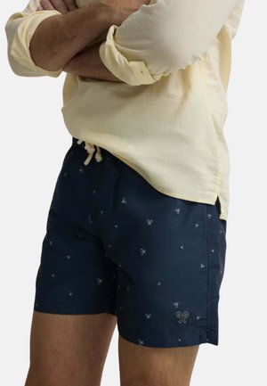 Hombre con bañador corto azul marino con pequeños estampados de raquetas de tenis y camisa de manga larga de color amarillo claro con las mangas remangadas.