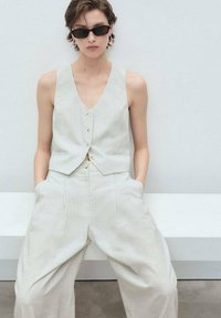 Lichtbeige, cropped vest met een V-hals en knoopsluiting, gecombineerd met wijde broek. Textured stof, minimalistisch ontwerp, stijlvolle zonnebril.