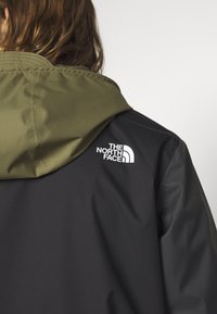 Chaqueta negra y verde oliva con capucha, que presenta un logo blanco en la parte posterior, textura suave y secciones de color contrastante.