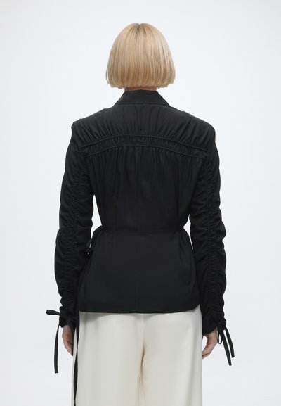 Personne aux cheveux blonds courts portant un haut noir froncé à manches longues et un pantalon blanc, vue de dos sur un fond uni.