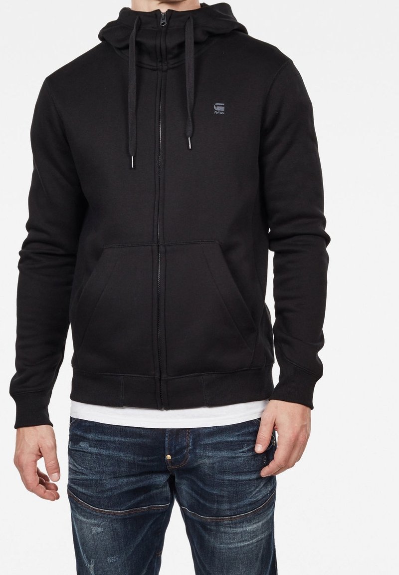 Felpa nera con zip realizzata in tessuto strutturato, due tasche frontali, cappuccio con stringhe e un piccolo logo sul petto. Abbinata a jeans blu.