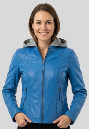Smiling woman wearing a blue leather jacket with a gray hooded sweatshirt underneath, standing against a plain light background.