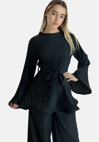 Haut noir plissé avec manches longues en cloche et taille nouée. Le tissu semble lisse avec une légère brillance, offrant une silhouette fluide.