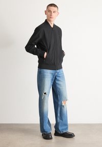 Black bomber jacket with a zip front and ribbed cuffs, paired with light blue distressed wide-leg jeans and black leather shoes.